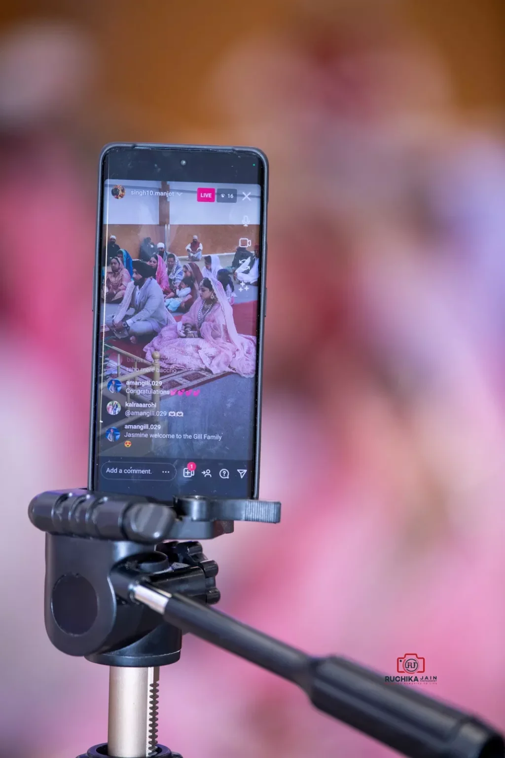 A smartphone on a tripod livestreaming a Sikh wedding ceremony, displaying a seated bride and groom in traditional attire