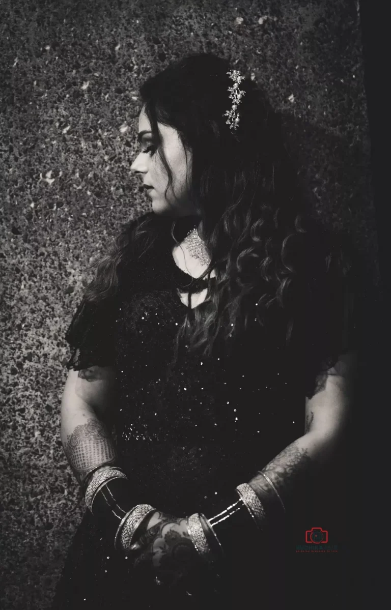 Black and white portrait of a woman in a dark outfit looking to the side with her hands clasped, standing against a textured wall