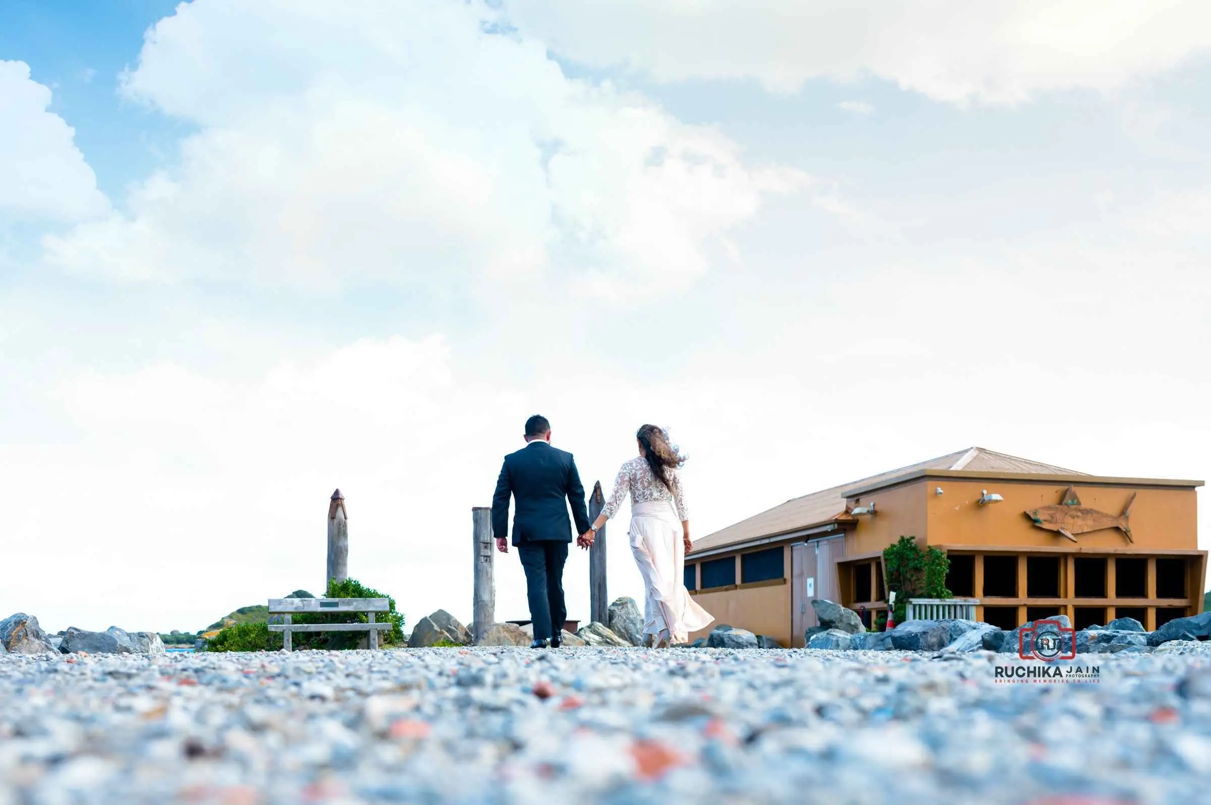 Bride and groom holding hands while walking along a rocky path outdoors, with a rustic building in the background