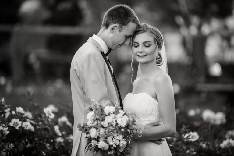 Bride and groom in a tender moment surrounded by flowers in a black and white photo.