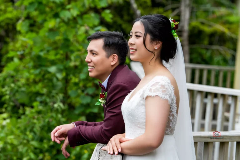 Bride and groom leaning on a wooden railing, smiling and enjoying a moment together outdoors