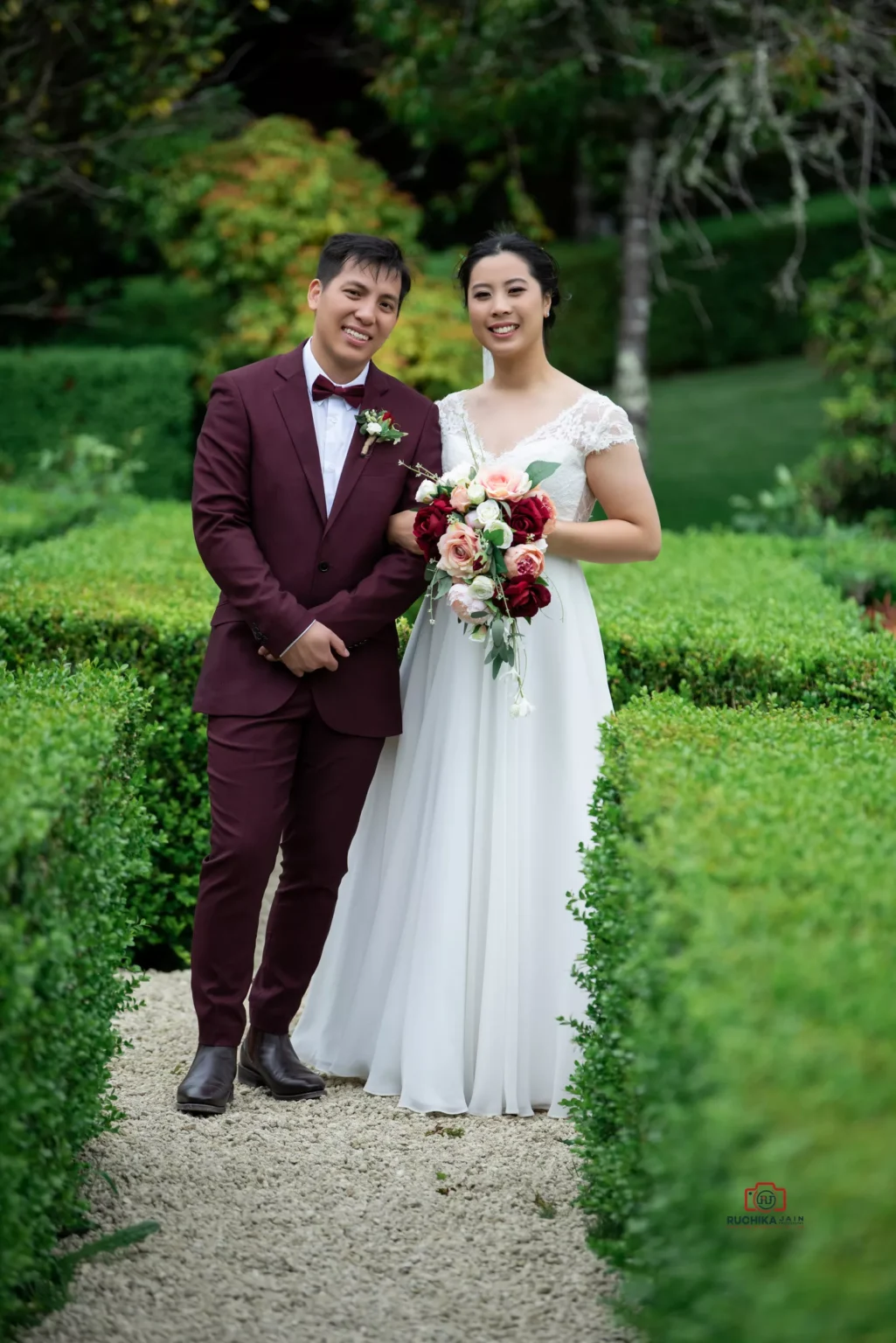 Bride and groom posing in a lush green garden pathway with the bride holding a bouquet of flowers