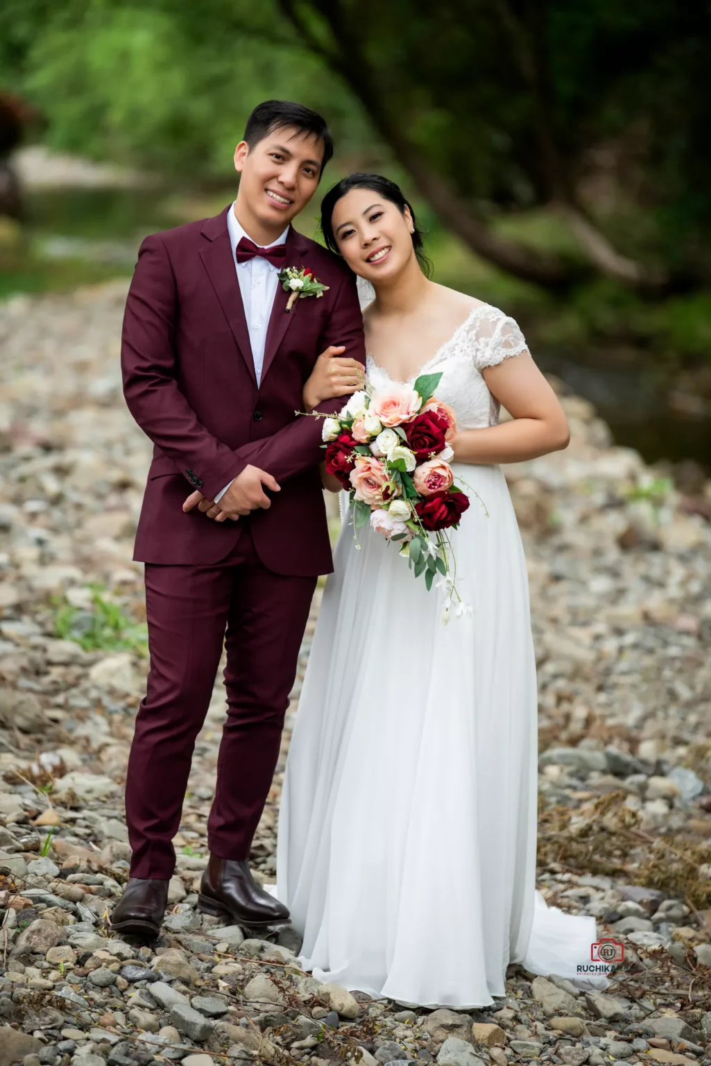 Bride and groom posing together on a rocky riverside, smiling and holding a bouquet.