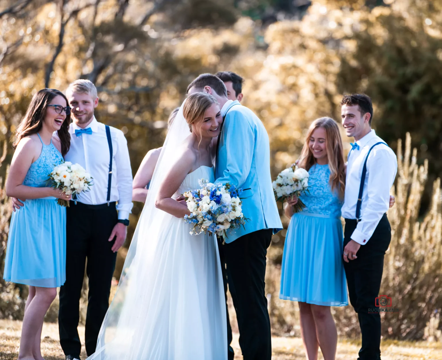 Bride and groom sharing an embrace with bridesmaids and groomsmen smiling in the background during an outdoor wedding