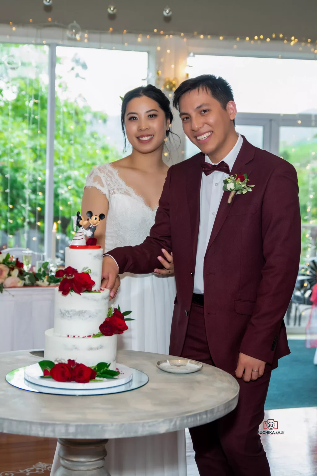 Bride and groom smiling as they cut a white wedding cake adorned with red roses and a cartoon topper