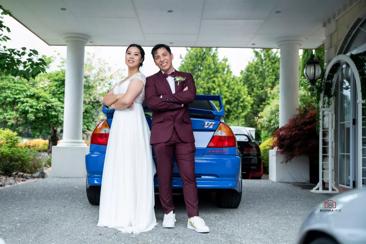 Bride and groom standing back-to-back in front of a blue car, smiling confidently on their wedding day