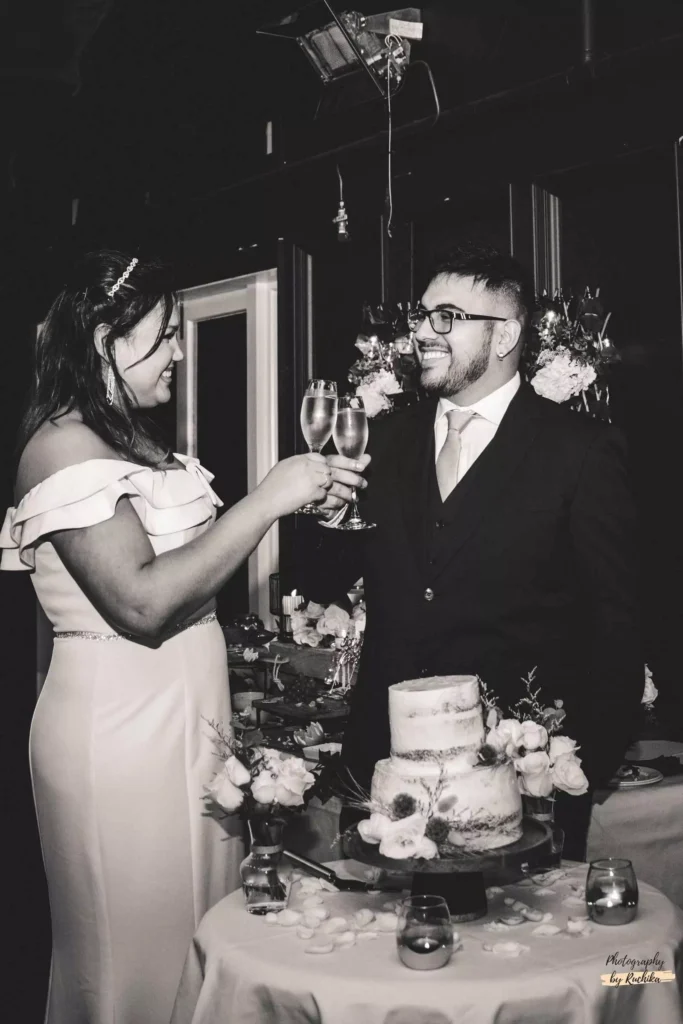 Bride and groom toasting with champagne glasses at their wedding reception, standing beside a decorated cake