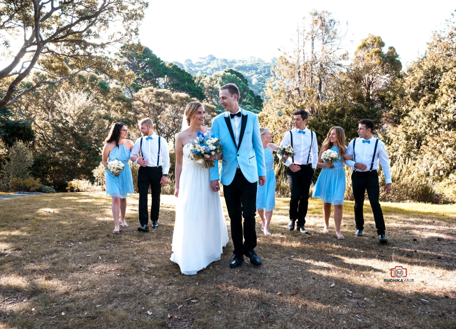 Bride and groom walking with their bridal party in a scenic outdoor setting, with bridesmaids in blue dresses and groomsmen in suspenders