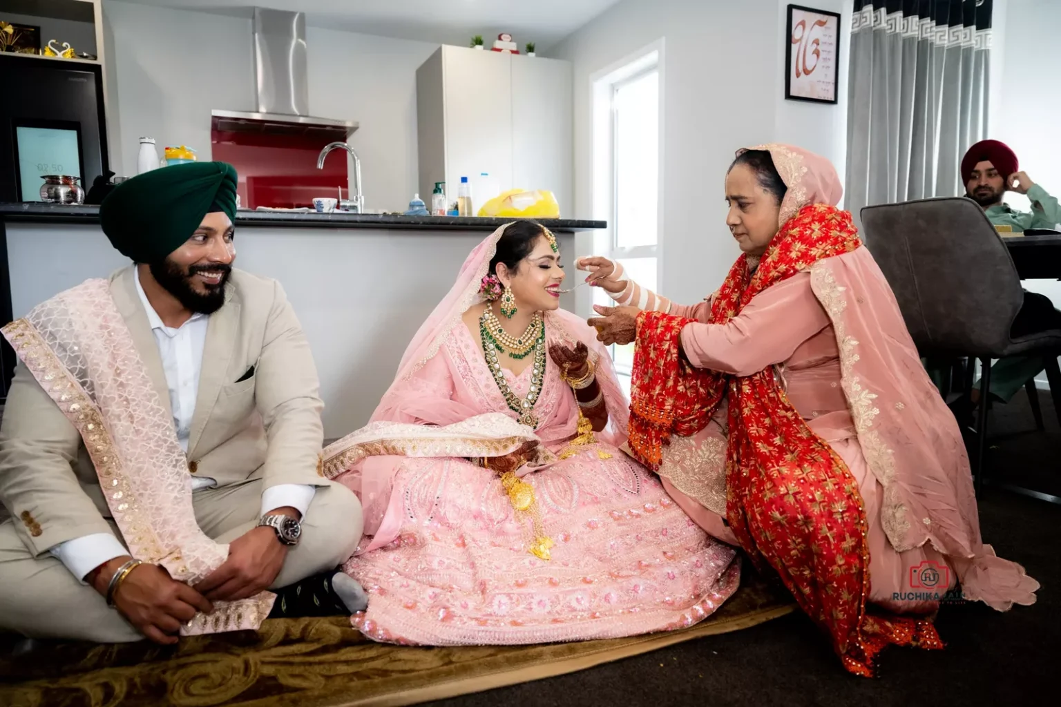 Bride being fed a sweet by a family member while sitting next to the groom in a home setting