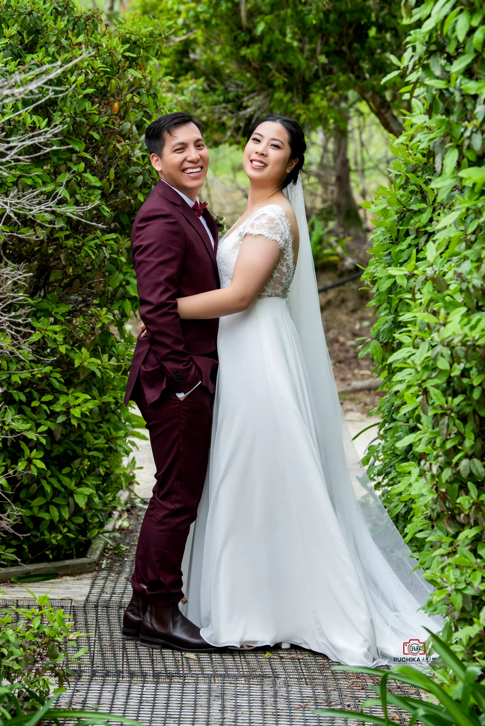 A joyful bride in a flowing white gown with lace sleeves and a long veil embraces the groom, dressed in a maroon suit with a matching bow tie, in a vibrant garden pathway surrounded by greenery.