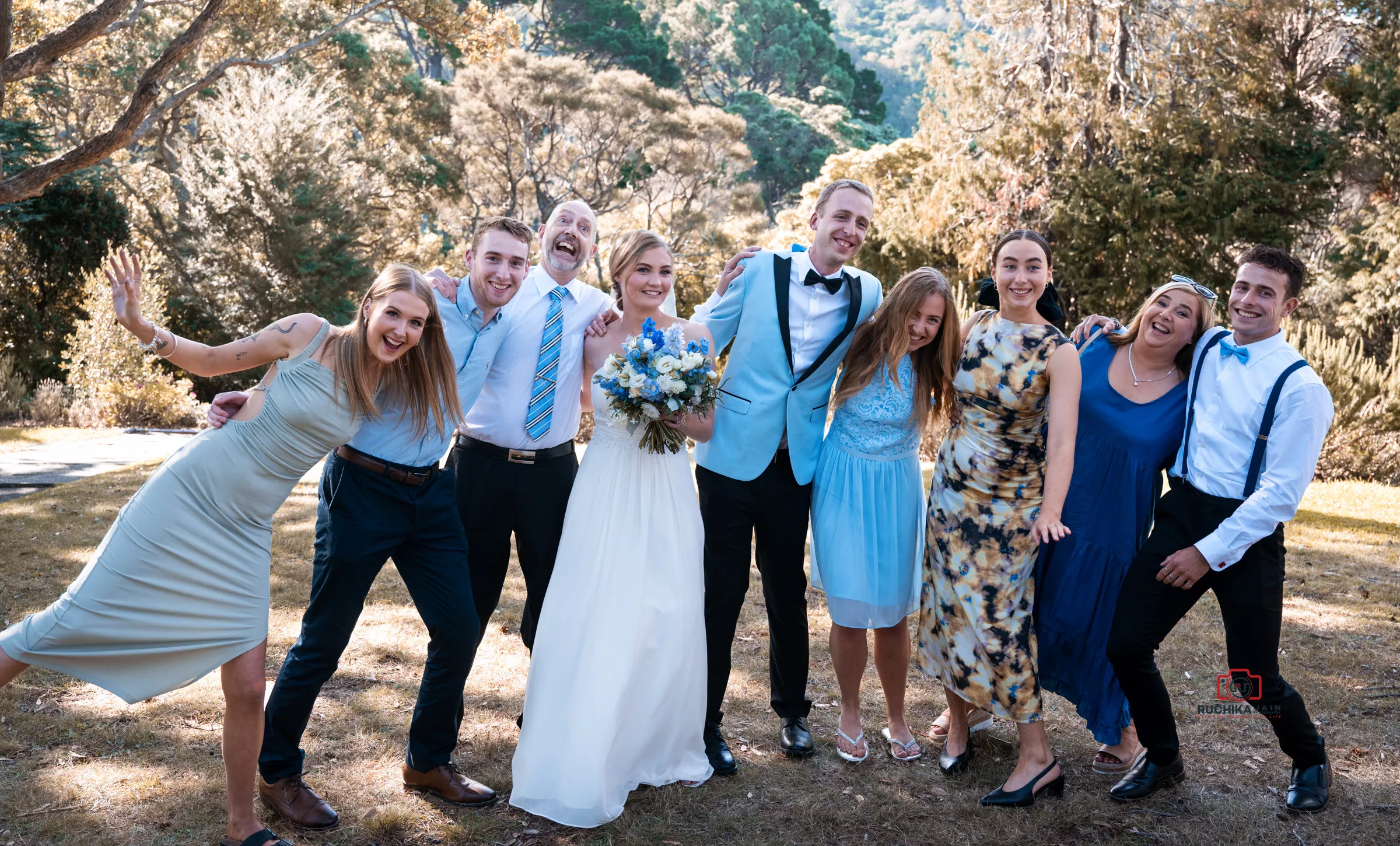 The bride and groom pose happily with their wedding party in an outdoor garden setting. The bride is in a white gown holding a bouquet of blue and white flowers, while the groom is in a light blue blazer and black bow tie. The group around them is dressed in coordinated outfits, smiling and sharing a lively moment.