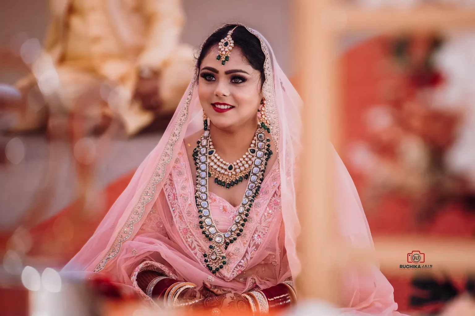 Bride in a pink lehenga sitting during a Sikh wedding ceremony, adorned with jewelry and smiling