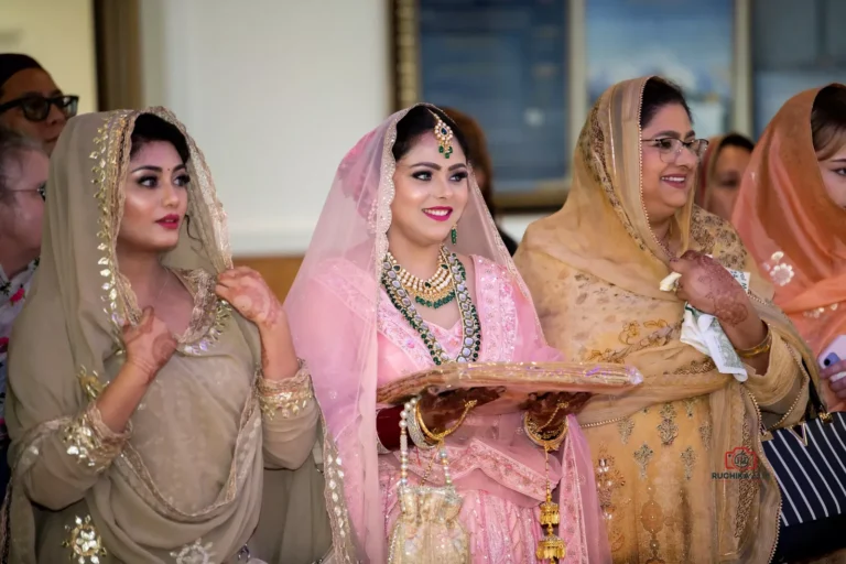 Bride in pink lehenga holding a ceremonial tray, standing with women in traditional attire during a Sikh wedding