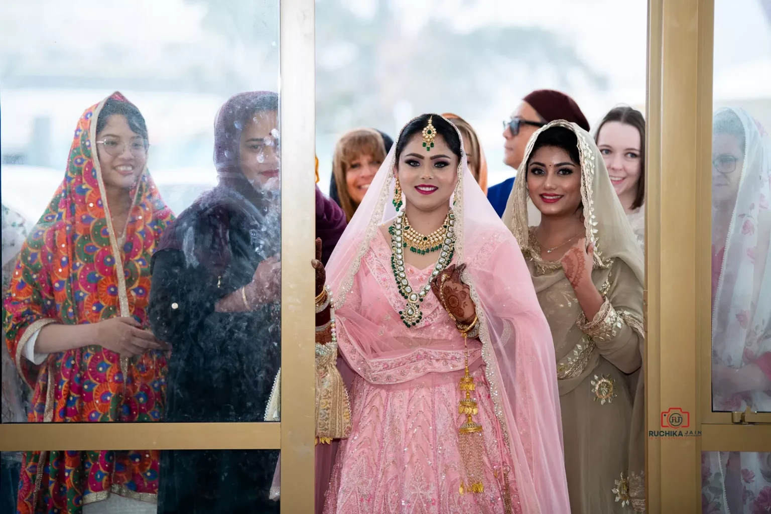 Bride in pink lehenga smiling with bridesmaids and guests through a glass door at a Sikh wedding