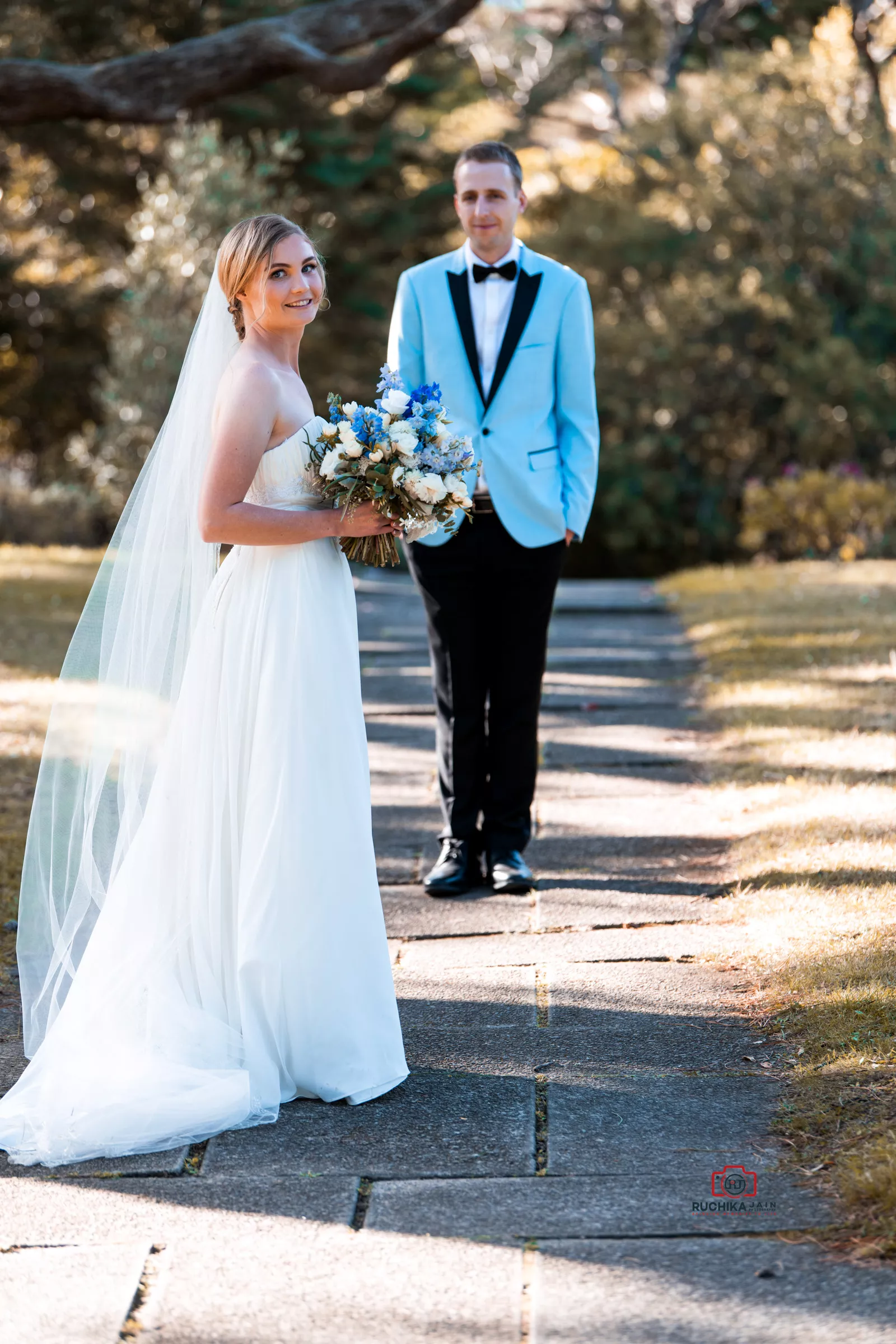 Bride in white gown holding a blue and white bouquet with groom in background wearing a light blue tuxedo jacket