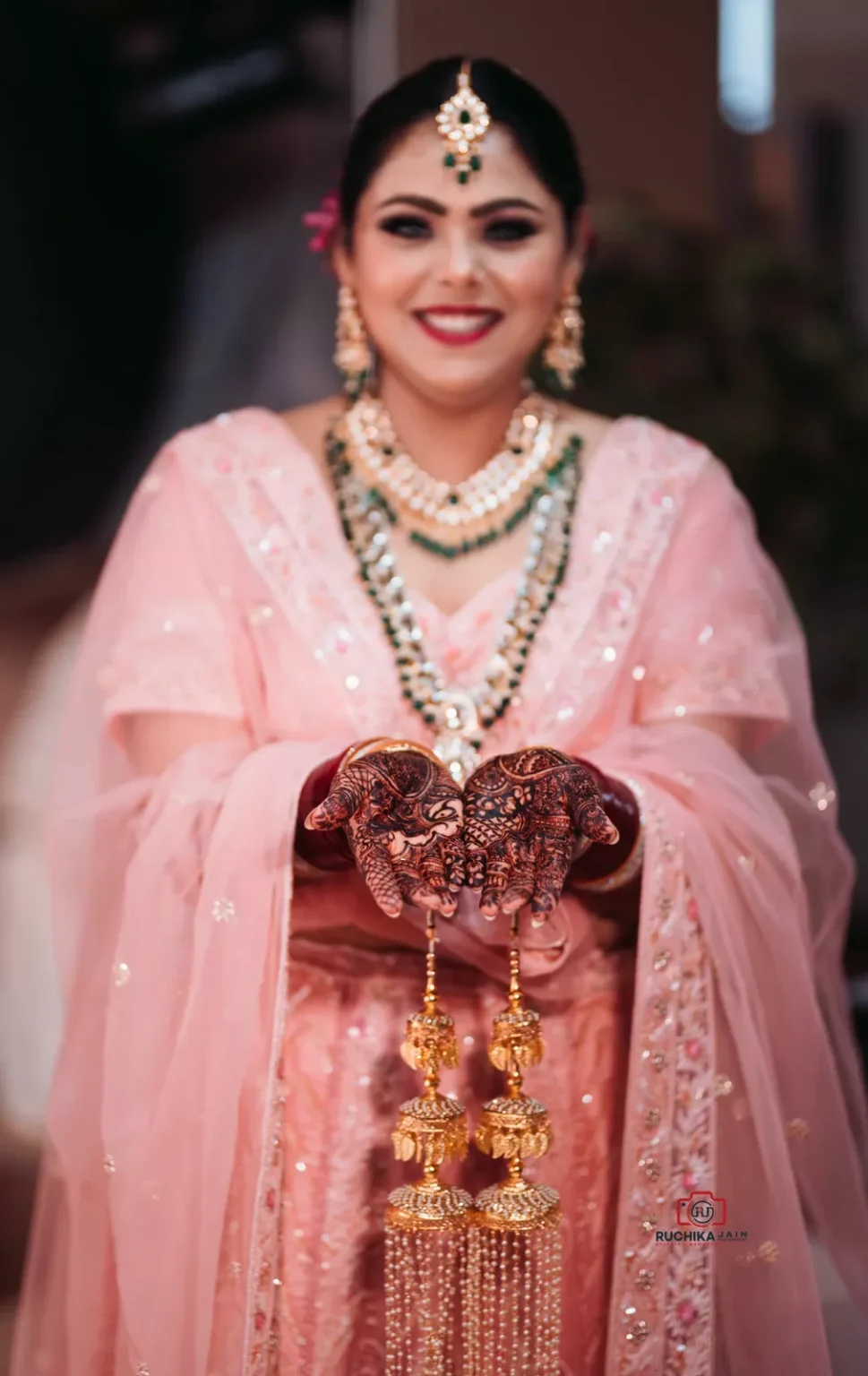 Bride showing intricate mehndi designs on her hands while holding gold bridal kalire in a pink wedding outfit