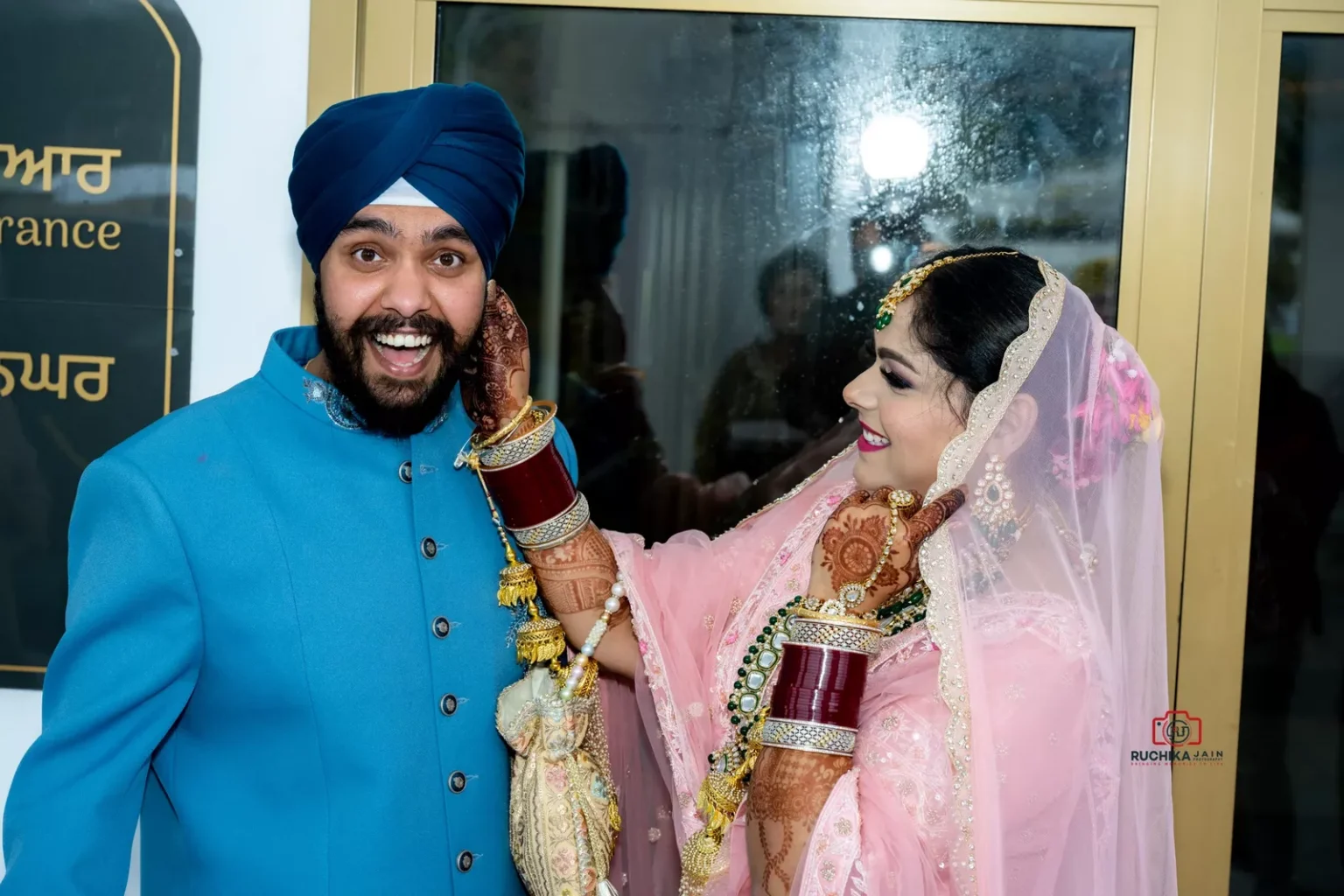 Bride smiling and playfully holding the face of a happy guest in a blue outfit at a Sikh wedding