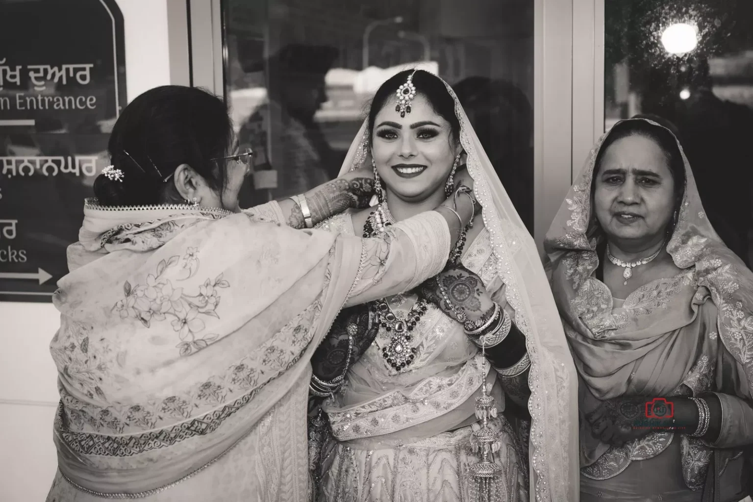 Bride smiling as family members adjust her jewelry in a black-and-white photo at a Sikh wedding