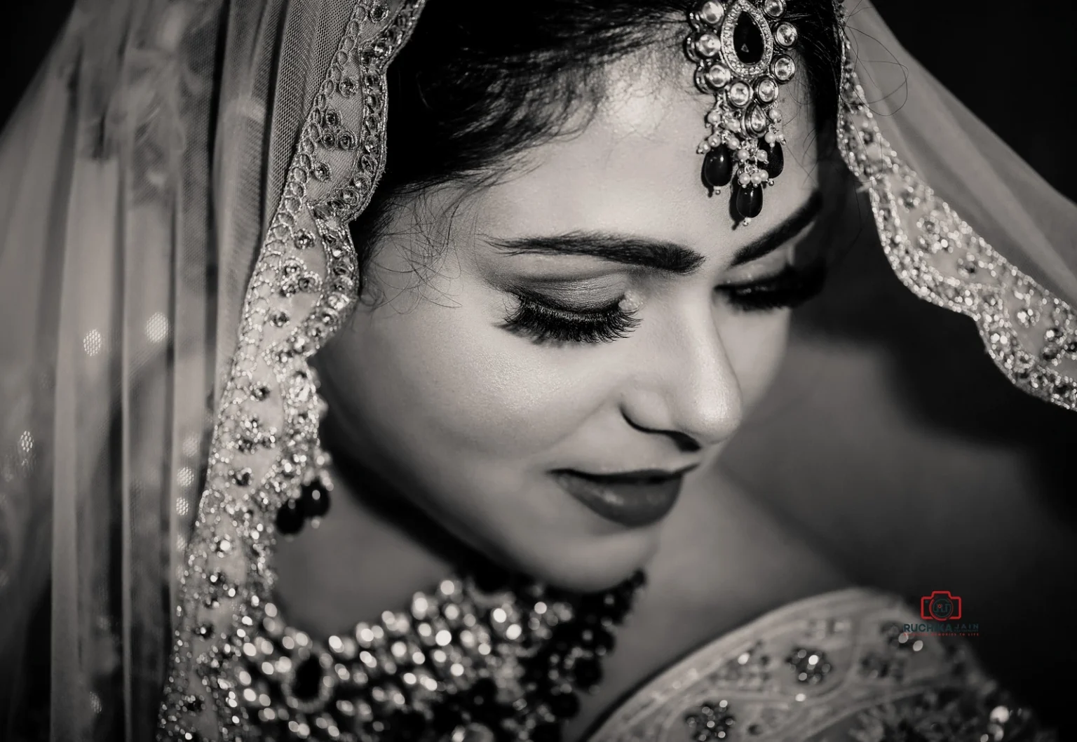 Close-up black and white portrait of a bride with traditional jewelry and veil, looking down with a soft expression