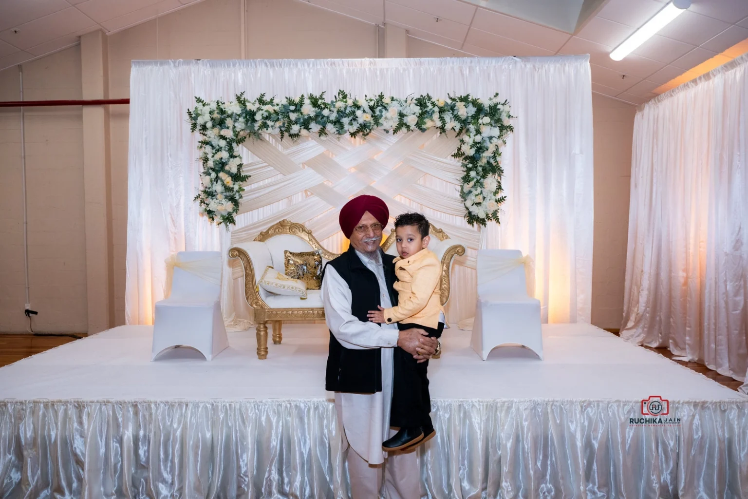 Elderly man holding a young boy in front of a decorated stage with white flowers and a gold couch