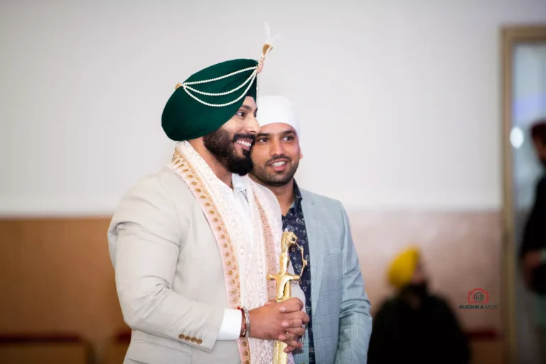 Groom in traditional Indian attire holding a sword and smiling, standing with a guest during a Sikh wedding ceremony