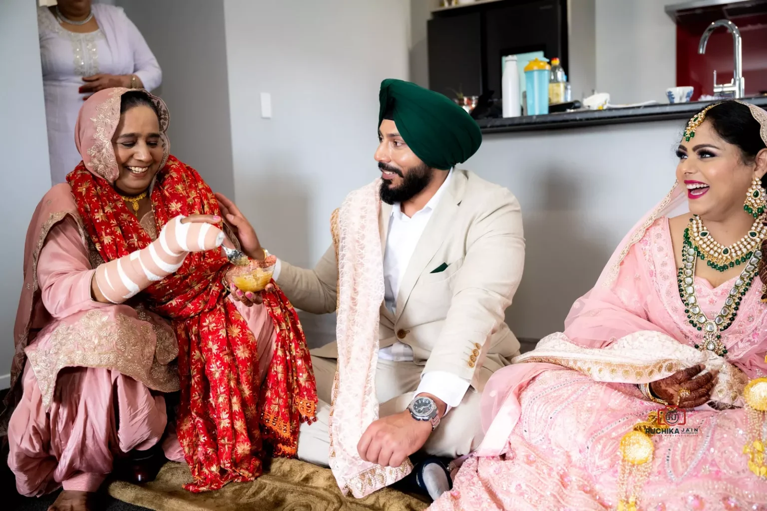 Groom smiling and sharing a light moment with a family member holding a bowl of sweets, while the bride laughs beside them.