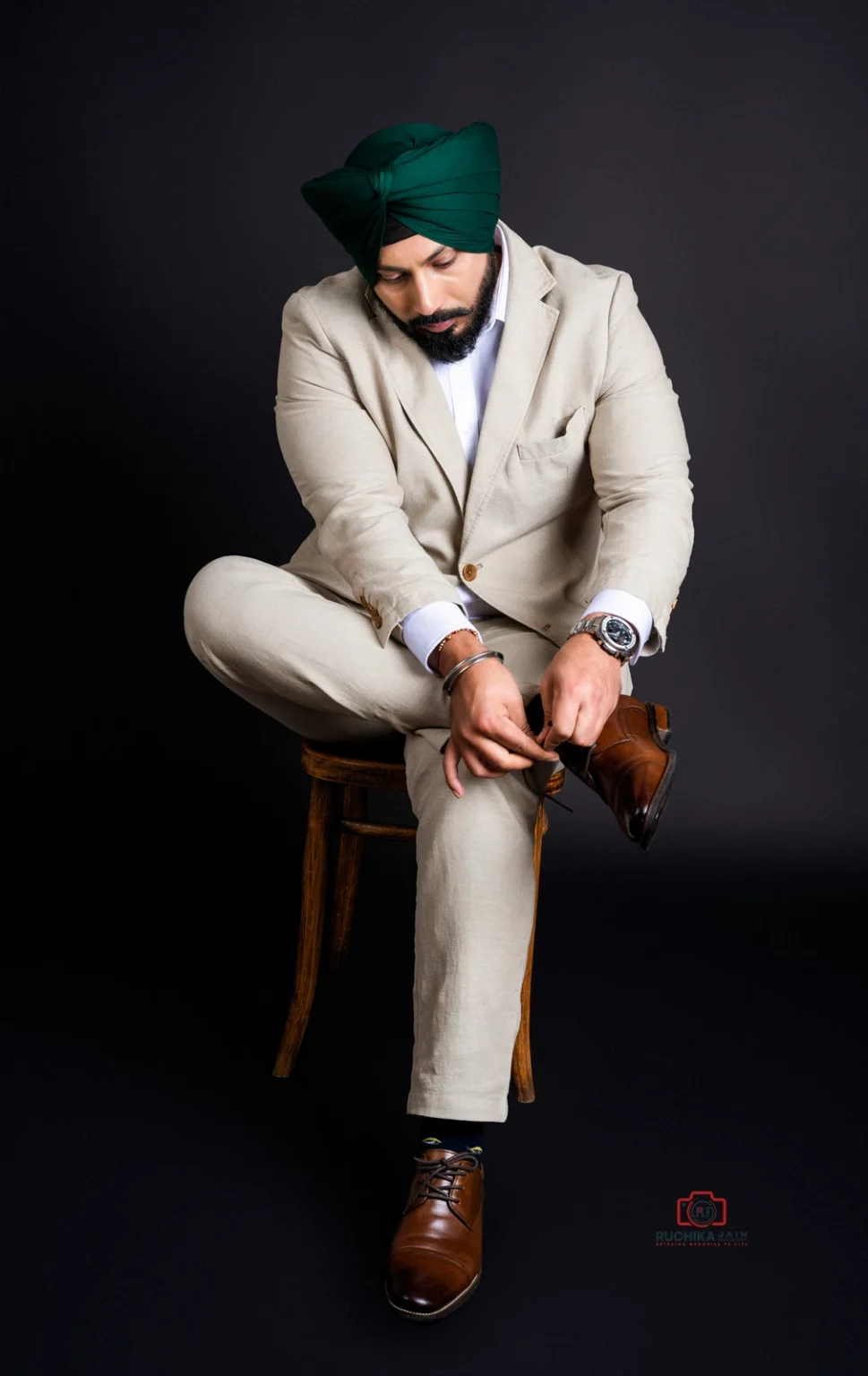 Man in a beige suit with a green turban sitting on a stool adjusting his brown leather shoes, against a dark background