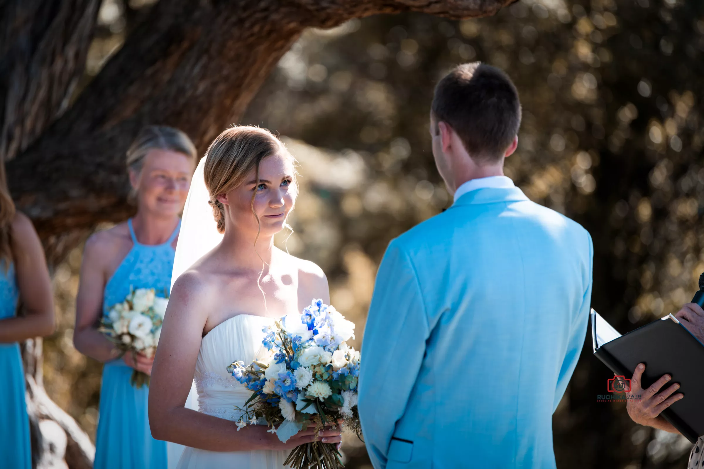 Bride in a strapless white gown holding a bouquet of blue and white flowers, gazing lovingly at her groom in a light blue suit during their outdoor wedding ceremony under a tree.