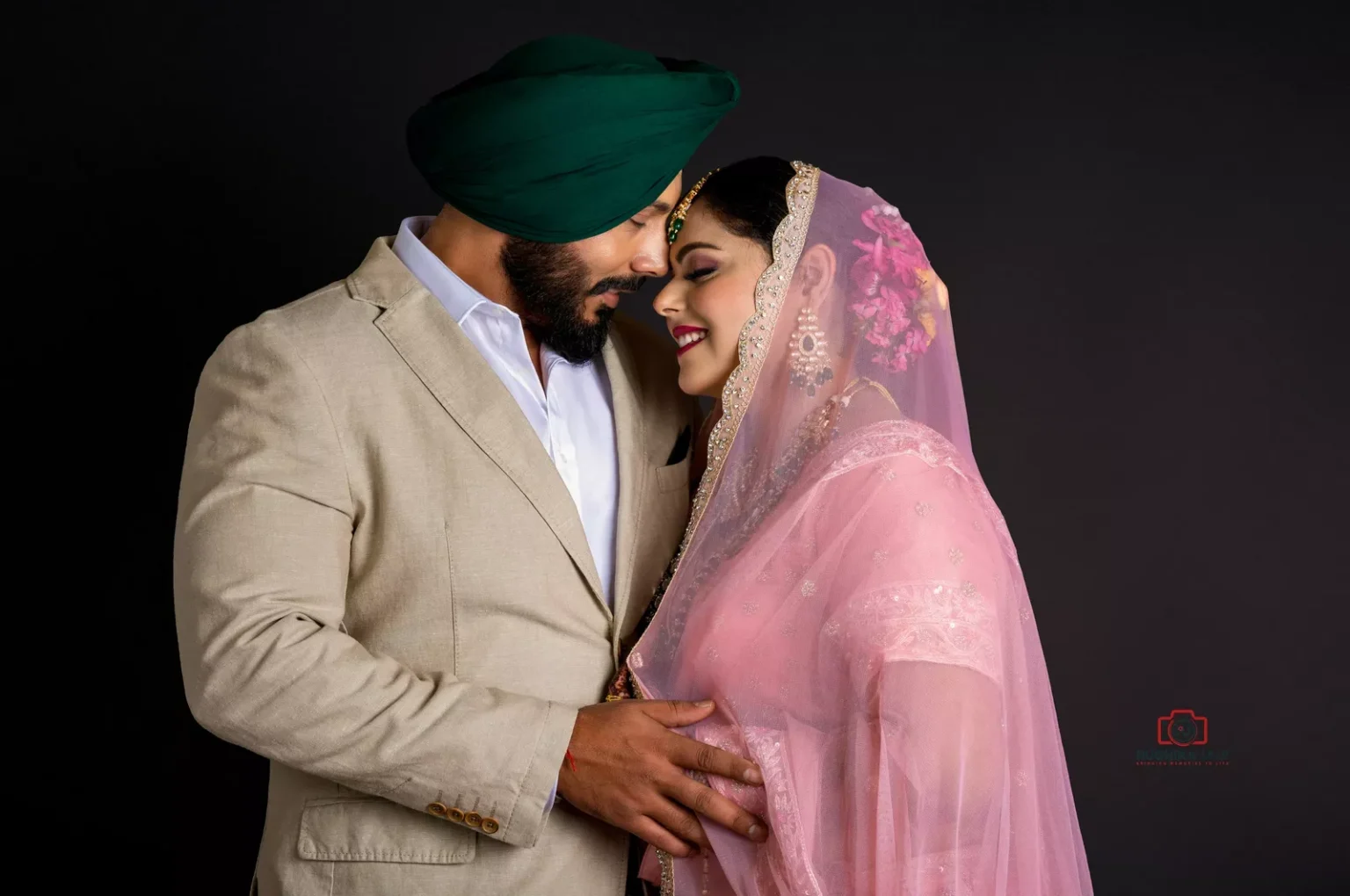 A Sikh couple sharing a tender moment, dressed in elegant traditional attire. The man wears a beige suit with a green turban, while the woman wears a delicate pink dupatta adorned with lace and floral details