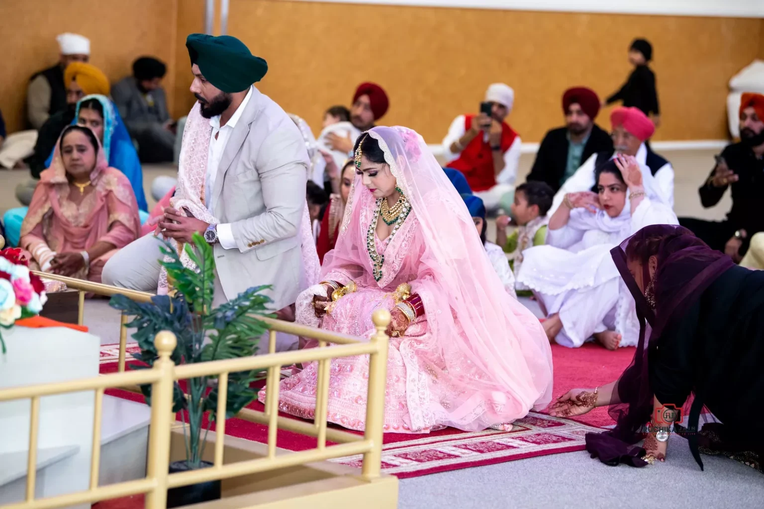 Sikh bride and groom kneeling during wedding ceremony in gurdwara with guests seated in the background