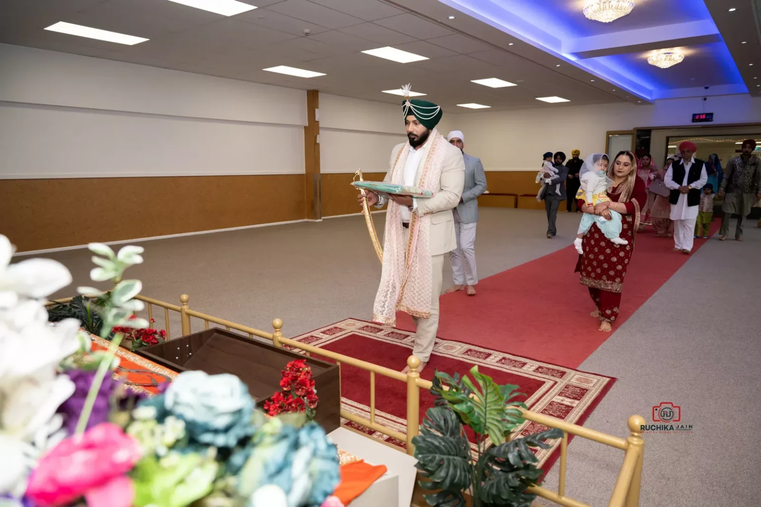 Sikh groom walking down a red carpet in a Gurdwara holding a religious book, with family and friends following