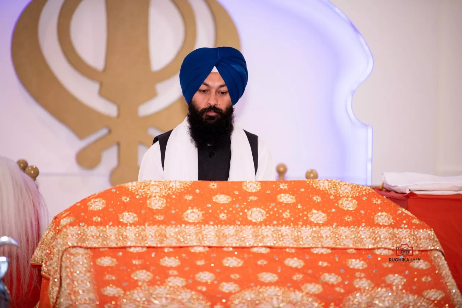 Sikh priest standing in front of a decorated Guru Granth Sahib covered in an orange cloth