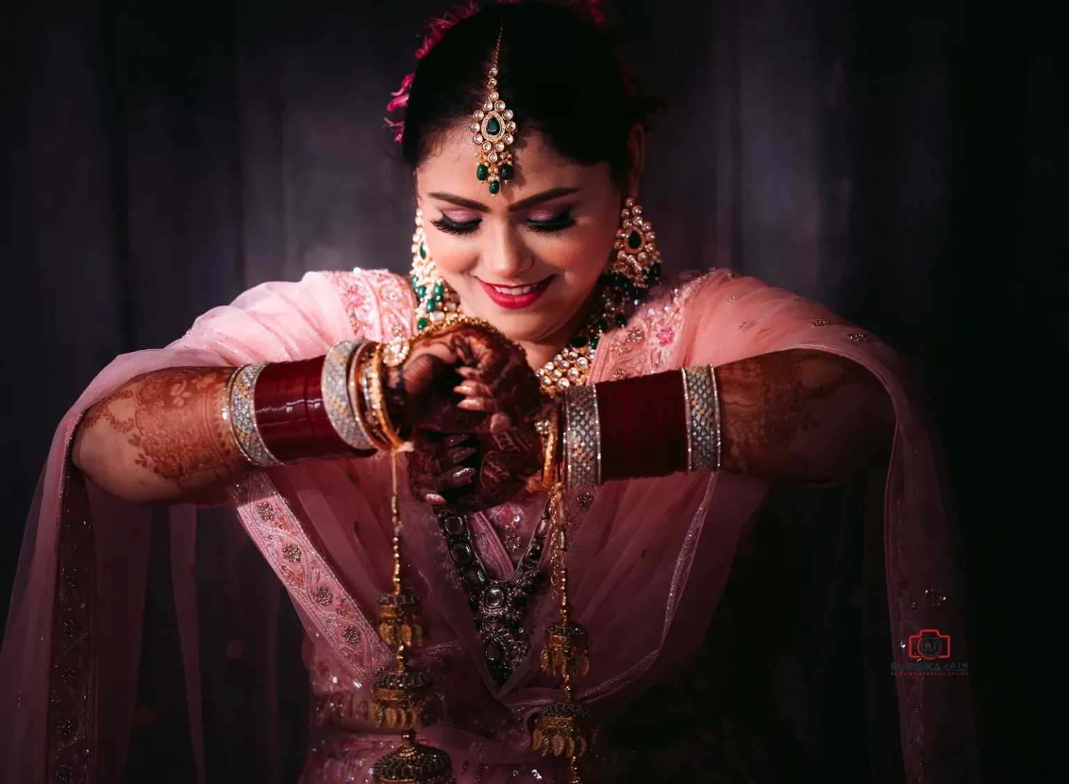 Smiling bride in a pink outfit looking down at her hands adorned with henna, bangles, and bridal jewelry, including kalire, under soft lighting