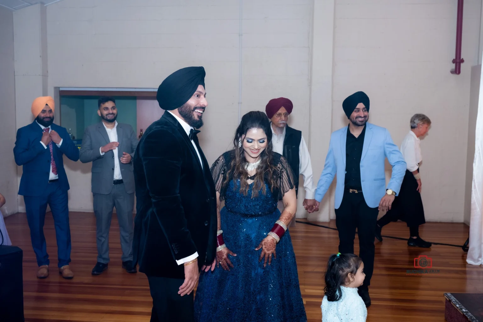 Smiling couple standing together on a dance floor at a celebration, surrounded by guests, including a young child in the foreground