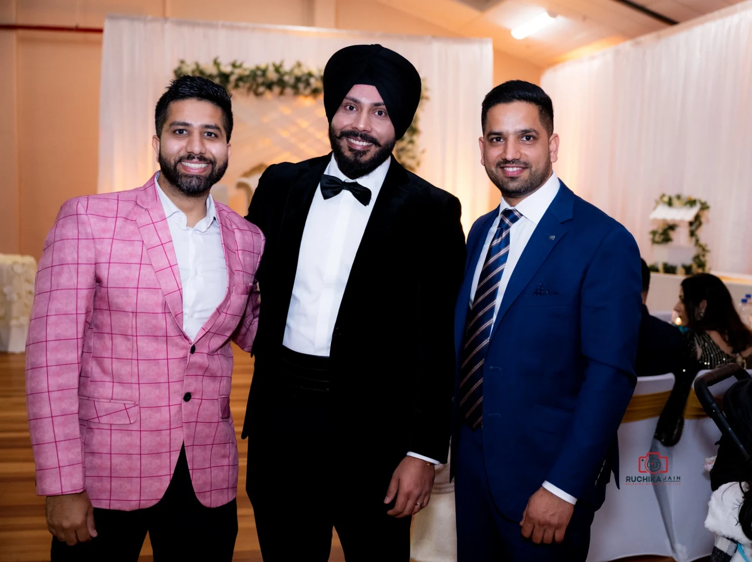 Three men dressed in formal attire standing together, smiling, with a decorative floral backdrop behind them