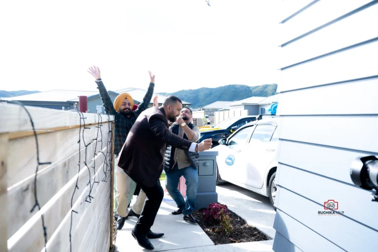 Three men outside a house, with one man in a brown blazer dancing while others cheer with raised hands, next to parked cars