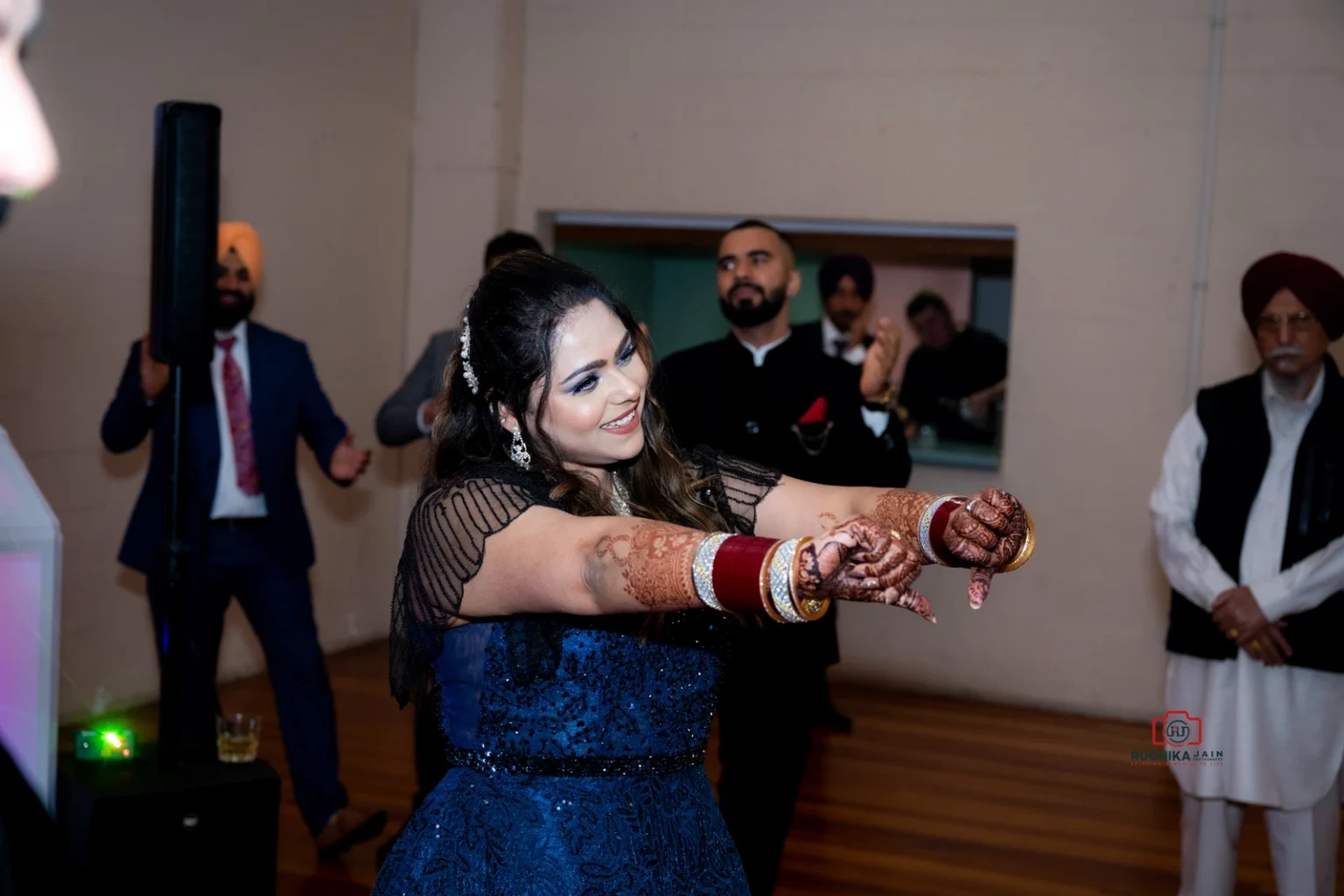 Woman in a blue dress with henna-decorated hands dancing at an indoor celebration, surrounded by guests