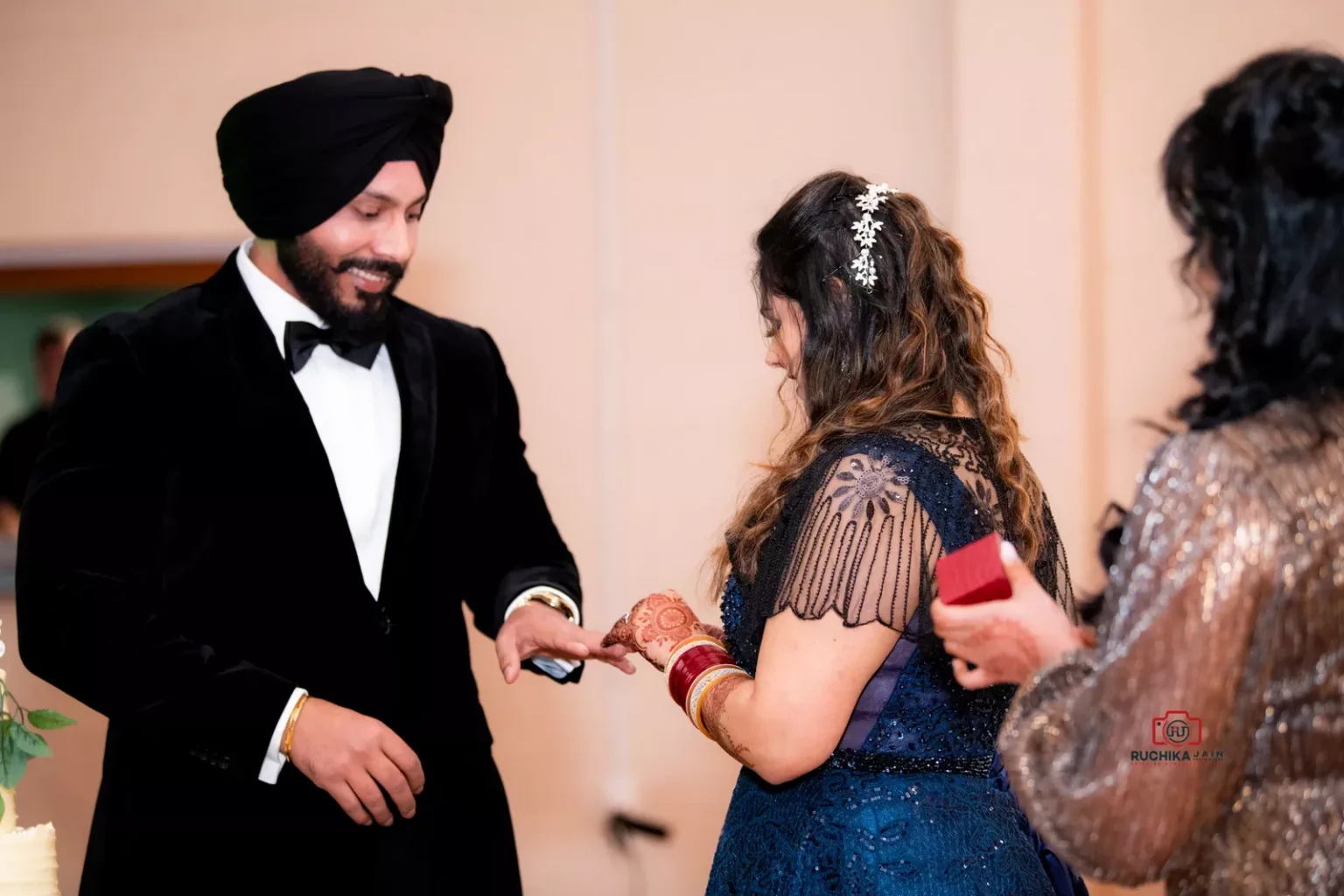 Woman placing a ring on a man's finger during a ceremony, with both dressed in formal attire. The man wears a black turban and tuxedo, and the woman is in a blue gown with henna on her hands