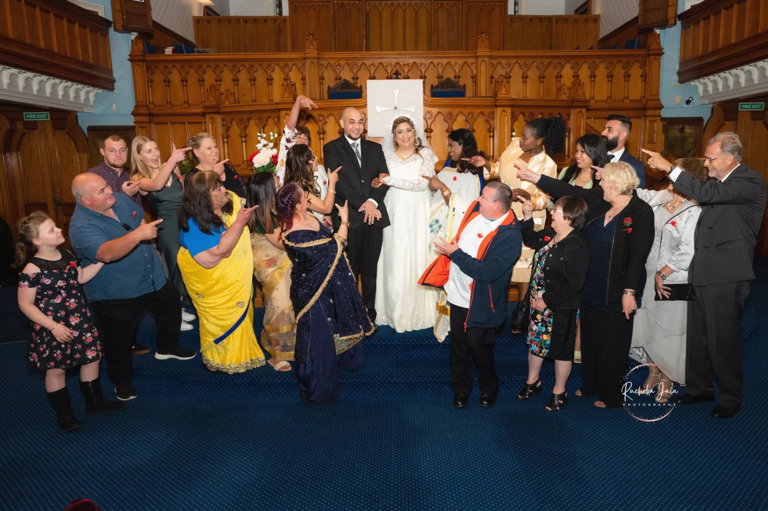 Anjali and Solomon surrounded by family and friends in a joyous group photo. The couple stands at the center of the image in wedding attire, with everyone pointing toward them in celebration. The background features a beautiful church setting with wooden accents.