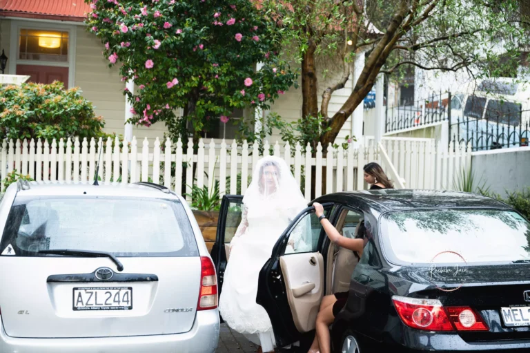 Bride in a white dress and veil stepping out of a car in front of a picket fence, with flowering trees in the background