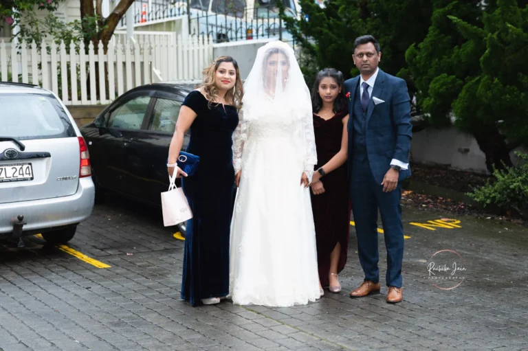 A bride in a white wedding gown with a veil, surrounded by her family, including a man in a navy suit, a young girl in a burgundy dress, and a woman in a dark velvet gown, standing outdoors near parked cars on a rainy day.