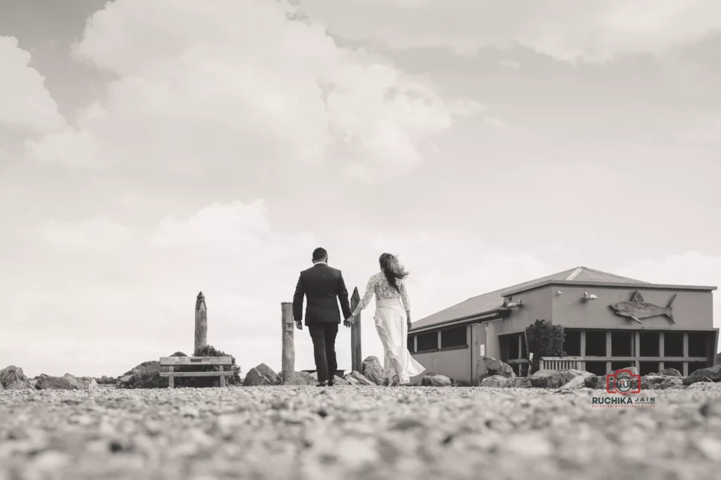 Black-and-white photo of a bride and groom holding hands as they walk towards the horizon by a coastal venue