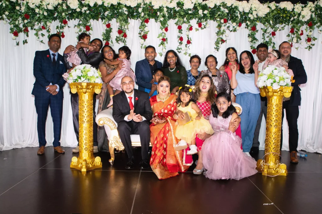 Group photo of the bride and groom surrounded by family and friends on a decorated wedding stage with floral backdrop and golden pillars