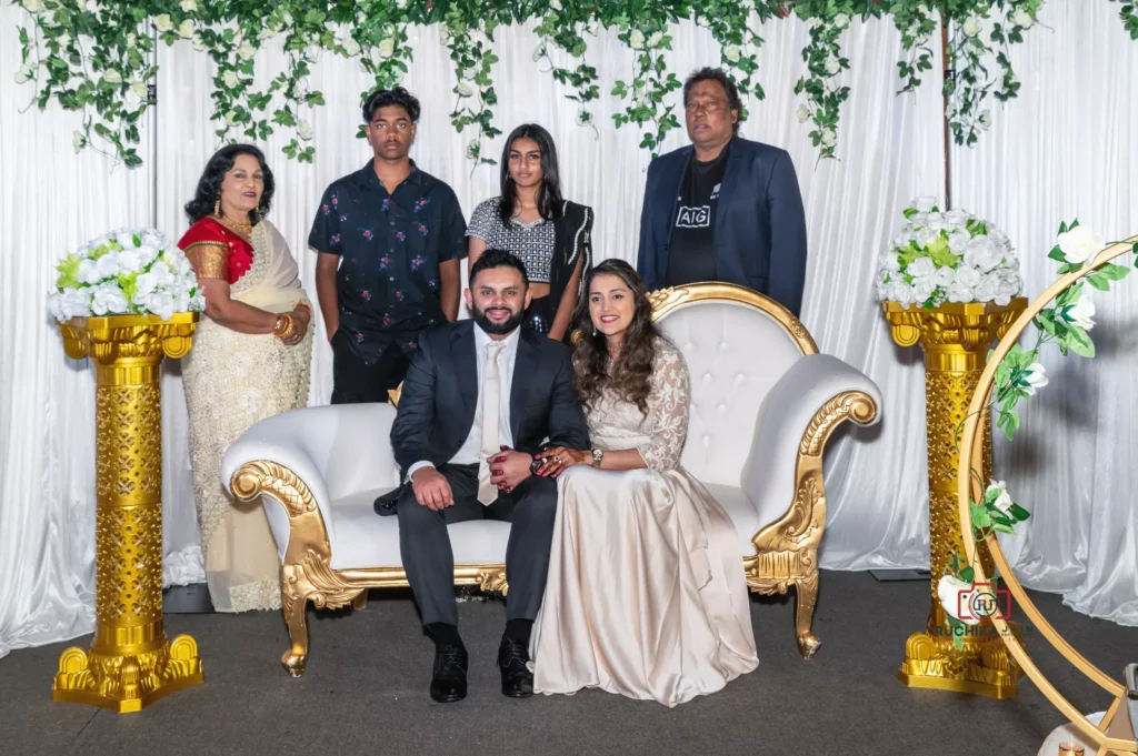 Bride and groom seated on a white and gold couch with family members standing behind, at a wedding reception decorated with green foliage and gold pillars