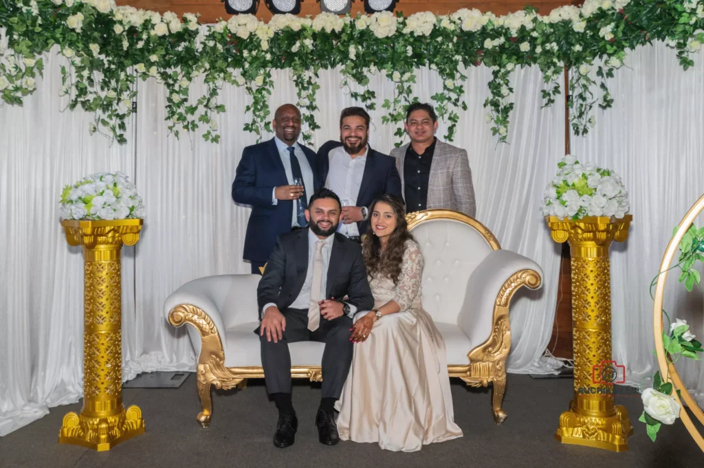 Bride and groom seated on a white and gold couch with guests standing behind, surrounded by floral and gold decor at a Wellington wedding reception