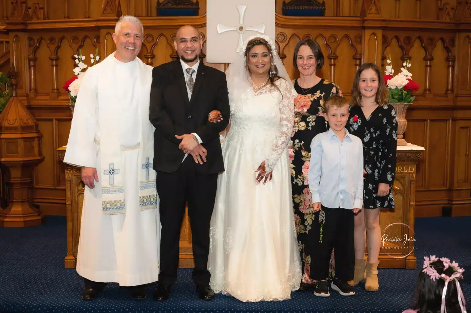 Bride and groom pose with the priest and family members inside a church, standing in front of a wooden altar