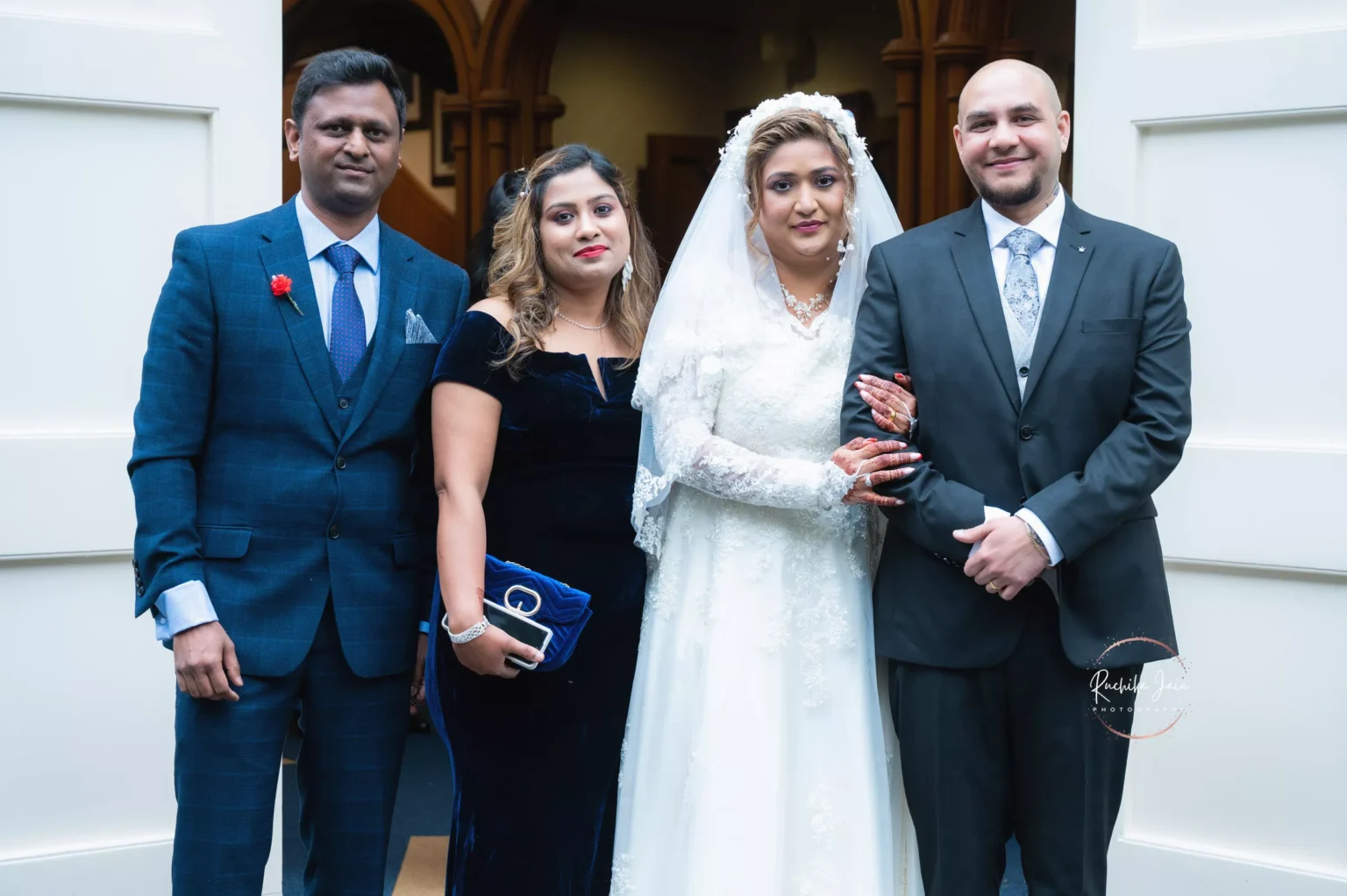 Bride in a white dress and groom in a suit standing with two guests in formal attire, posing together at the wedding venue entrance