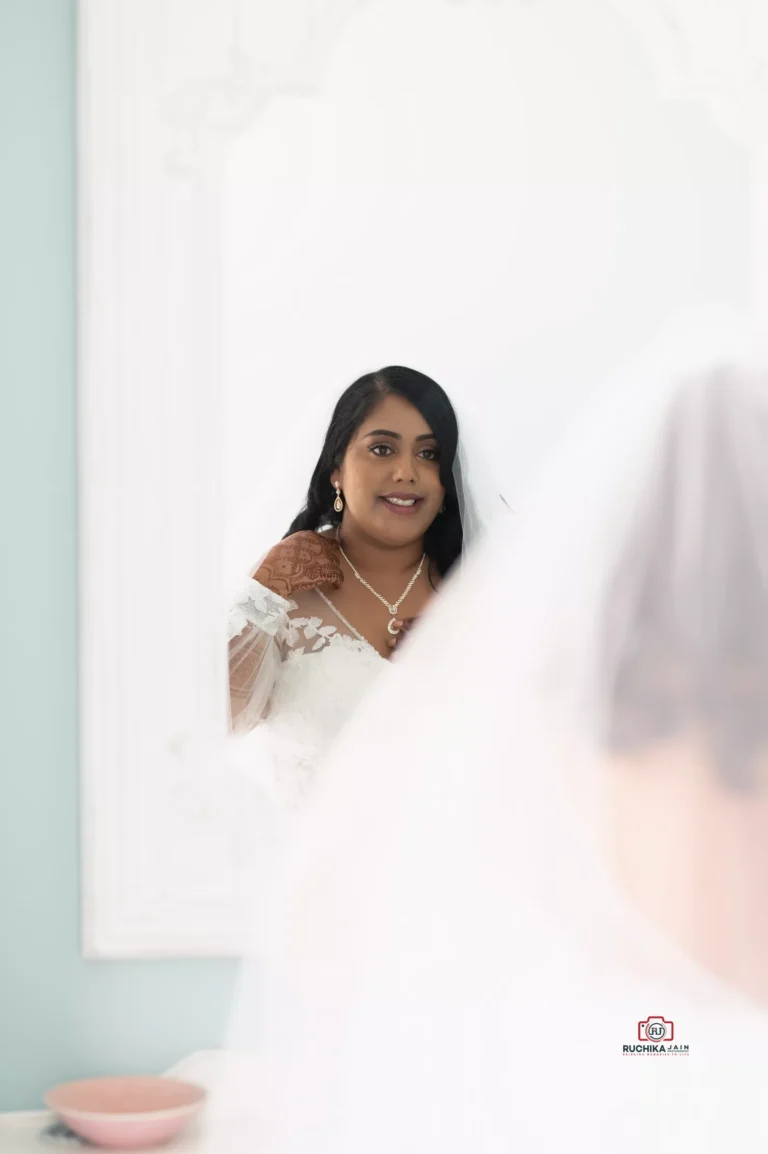 Bride looking into a mirror with a calm expression, wearing bridal jewelry and mehndi on her hand, captured in a soft, elegant setting