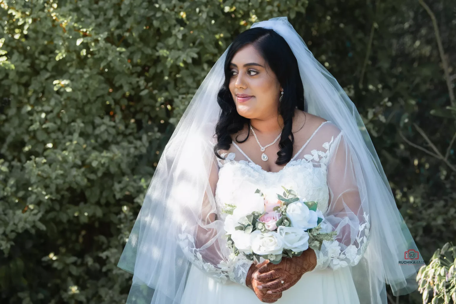 Bride standing outdoors holding a floral bouquet, wearing a white wedding gown with a lace veil, surrounded by lush greenery