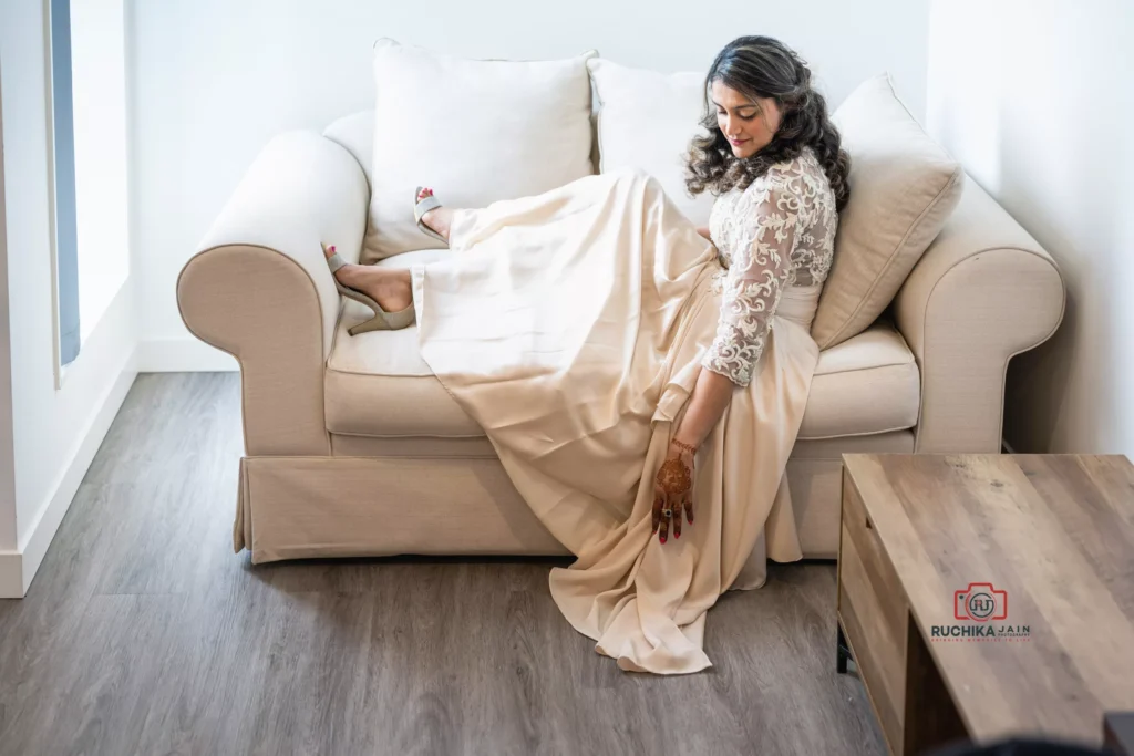 Bride in an elegant lace gown seated on a sofa, adjusting her dress with a serene smile